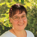 Porträt von einer Frau in weißem Shirt mit braunen Haaren und einer Brille.